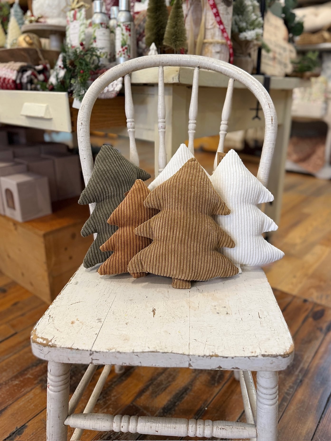 Set of Corduroy Stuffed Trees pillows in evergreen, cookie, camel, and white arranged on a vintage white chair in a cozy holiday shop setting.