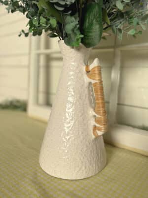 Footed off-white ceramic vase with curved rattan wrapped handles displayed with faux florals