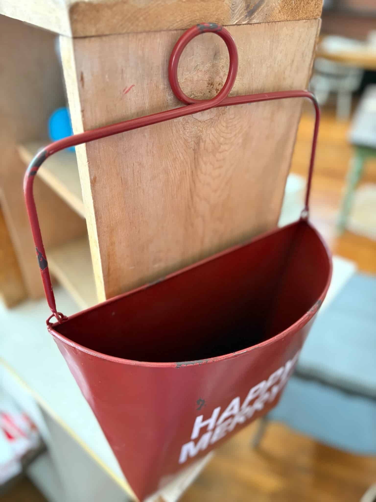 Distressed red metal wall bucket with curved handle and white “Happy Merry” stamped text.