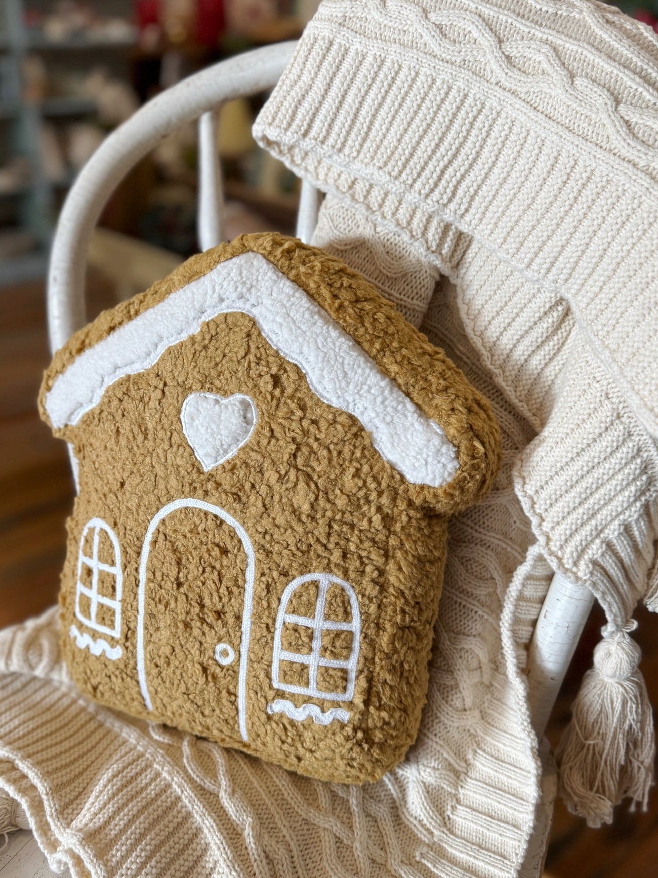 The gingerbread house pillow rests on a cream knit blanket draped over a white chair.