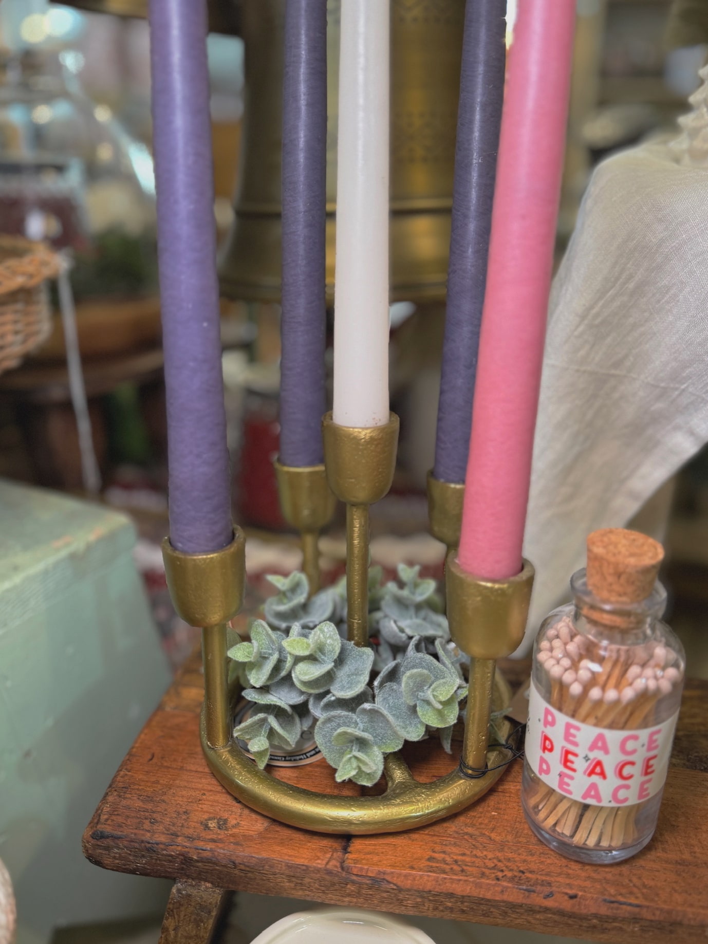 Gold Advent candle holder styled with purple, pink, and white taper candles, eucalyptus greenery, and a vintage match jar labeled “Peace.”