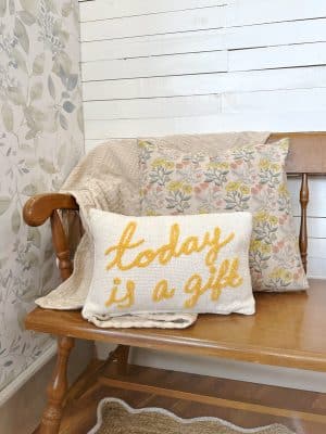 Cotton floral throw pillow, embroidered “Today is a Gift” lumbar pillow, and neutral waffle blanket styled on a wooden bench with floral wallpaper in the background.
