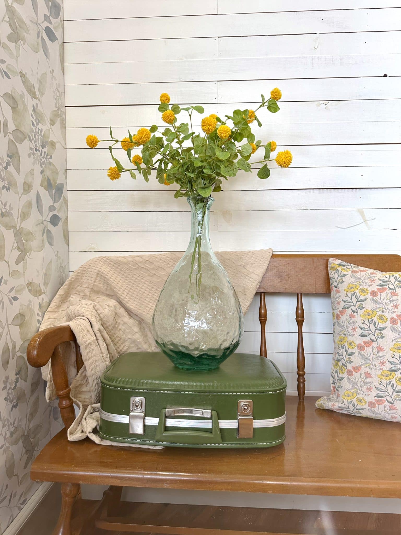 Large green bubbled glass vase with yellow faux florals styled on a vintage green suitcase, next to a neutral throw and floral pillow on a wooden bench.