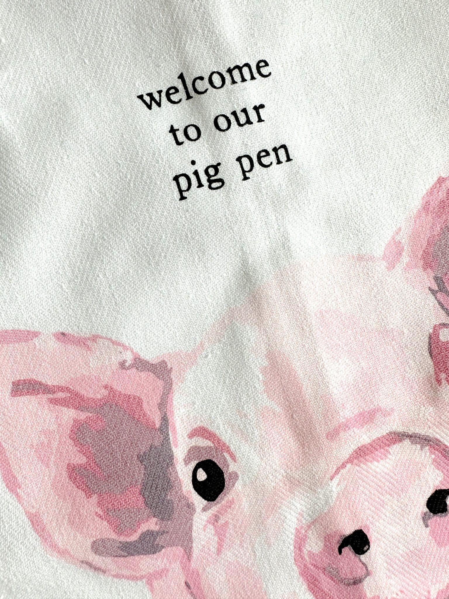 Close-up of pink watercolor pig illustration and “welcome to our pig pen” text on white cotton tea towel.