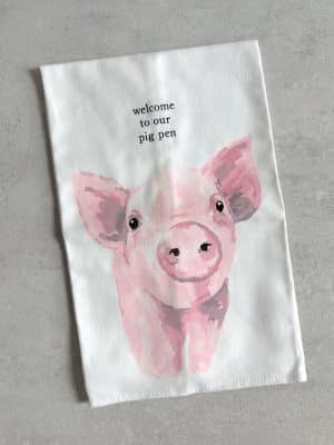 Cotton tea towel with a large pink watercolor pig and the phrase “welcome to our pig pen” printed above it.