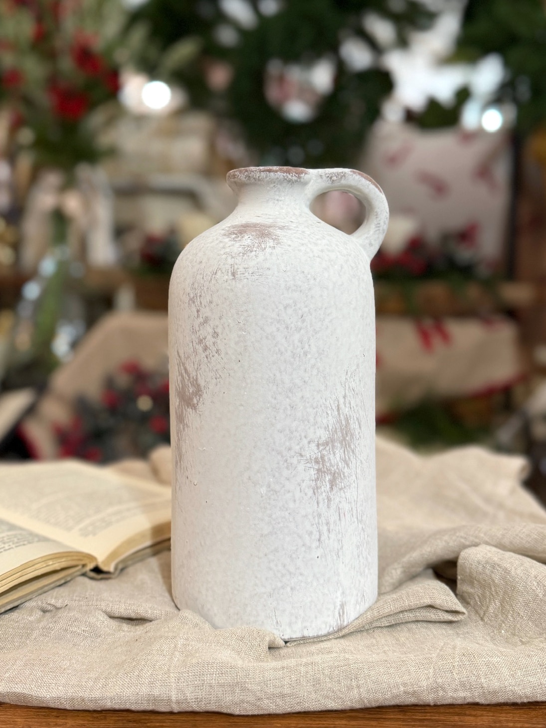 Rustic white single-handle vase displayed upright on a neutral cloth with soft holiday décor in the background.