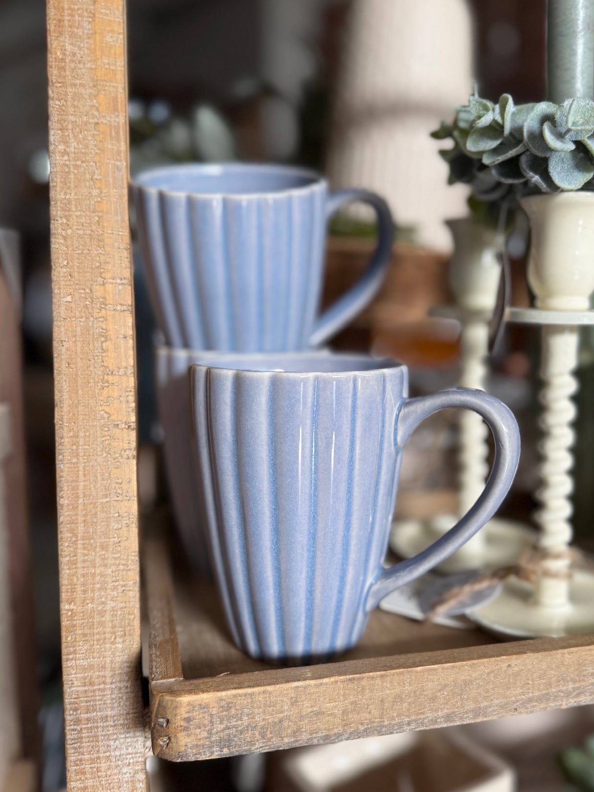 Set of stoneware ribbed mugs in light blue with reactive glaze finish, stacked on a wooden shelf.