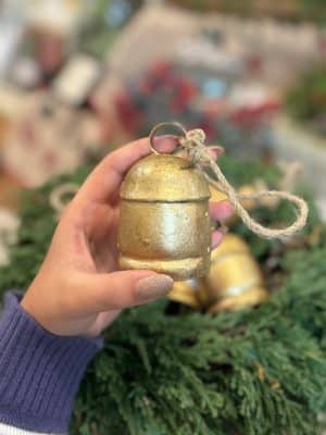 Hand holding a small distressed gold metal bell with jute rope hanger, styled with greenery and soft holiday decor in the background.