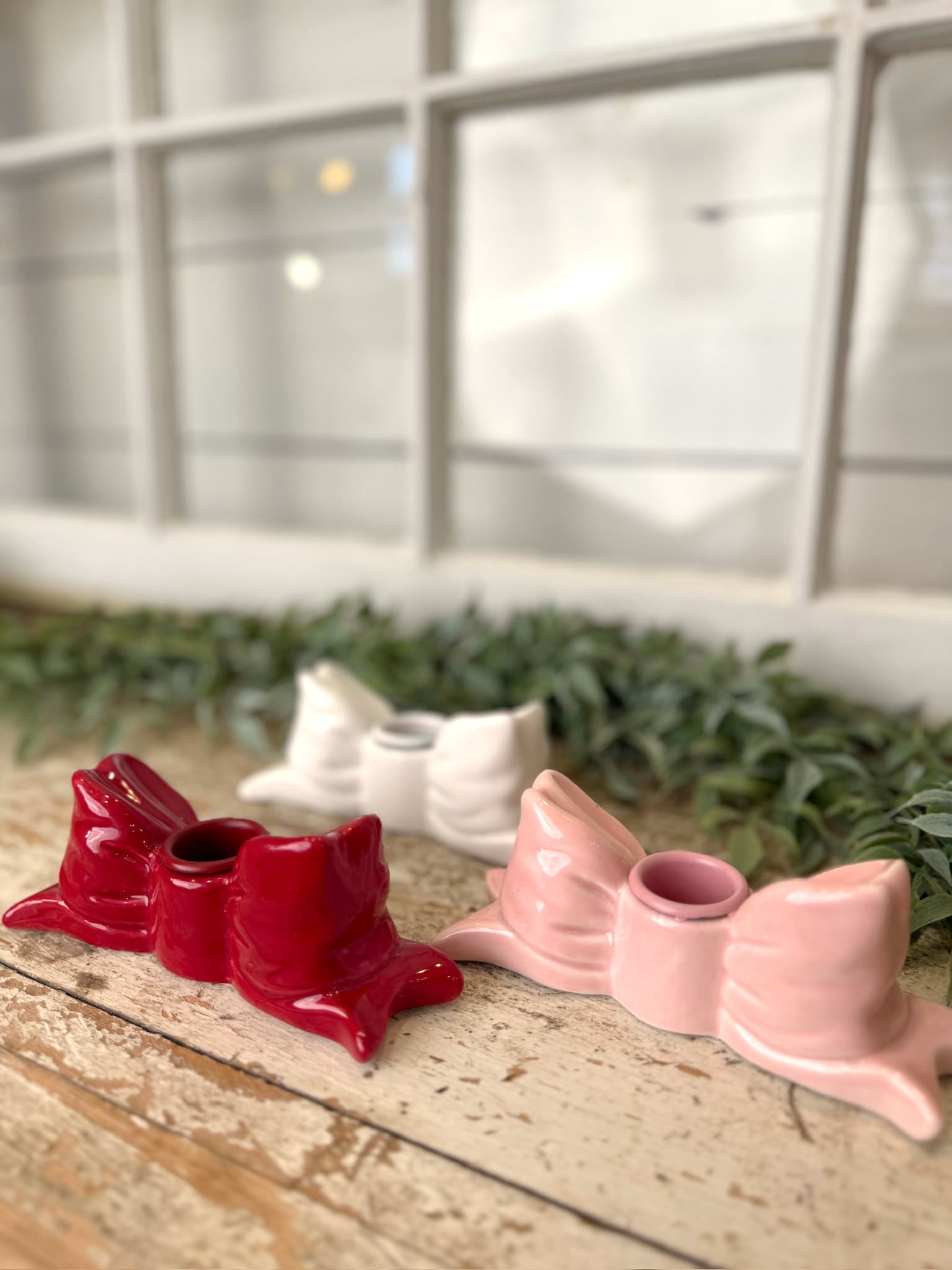 Pink, red, and white stoneware bow taper candle holders styled together on a rustic wood surface with soft greenery.