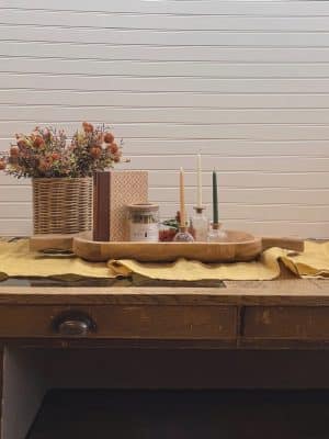 Styled fall vignette featuring a large crafted dough bowl with handles, stonewashed mustard table runner, wicker basket with orange pincushion picks, decorative vintage book set in neutral colors, Warm Abode deep teal matches, skinny taper glass candle holders, and assorted skinny taper candles.