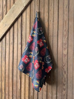 Black microfiber tea towel printed with red and gold Chiefs-themed designs including jerseys, footballs, heart sunglasses, and โKingdomโ pennants, hanging on a wooden wall.