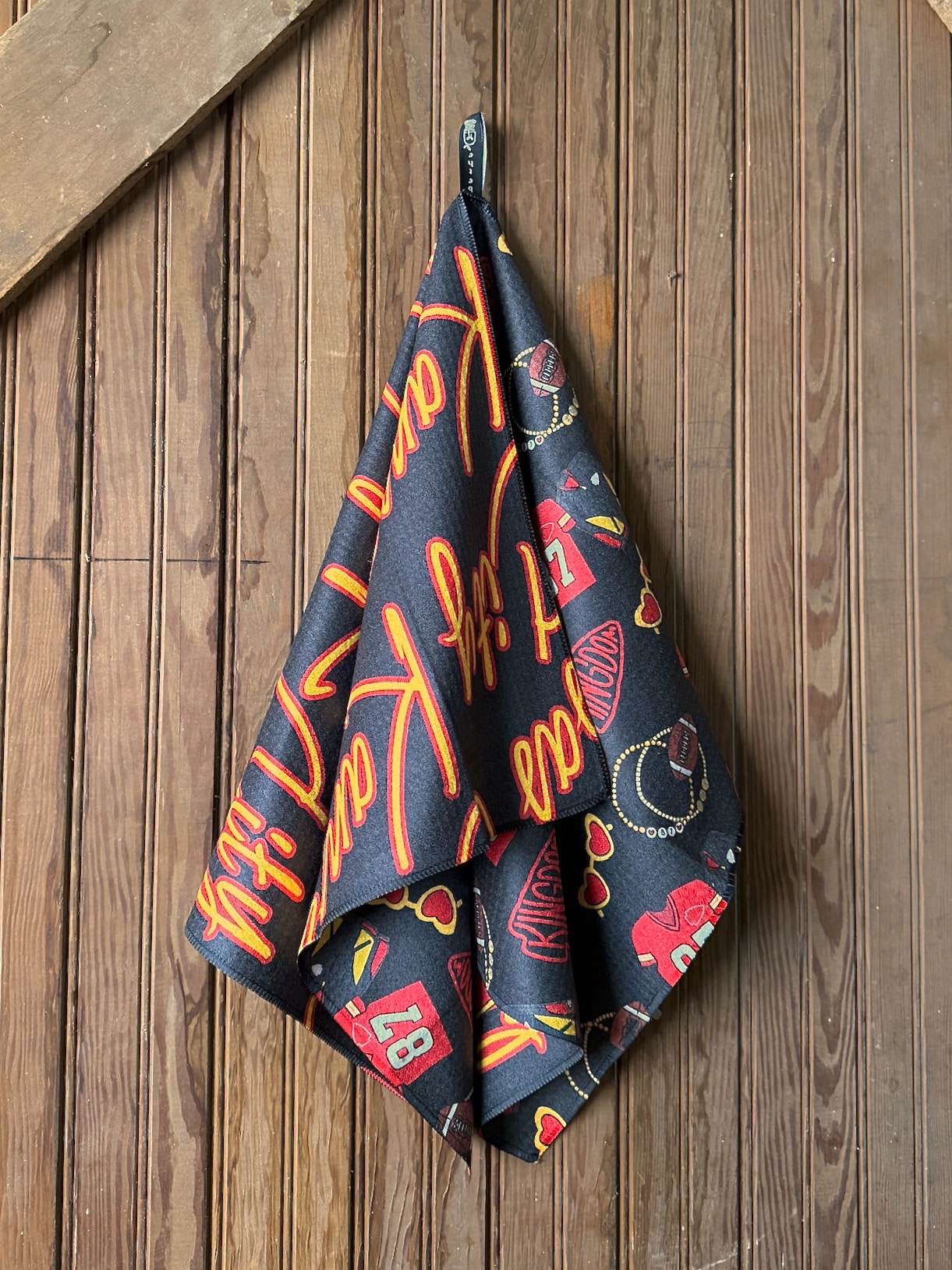 Black microfiber tea towel featuring red and gold Kansas Cityโthemed prints, including โ87โ jerseys, footballs, and heart sunglasses, hanging from a wooden wall hook.