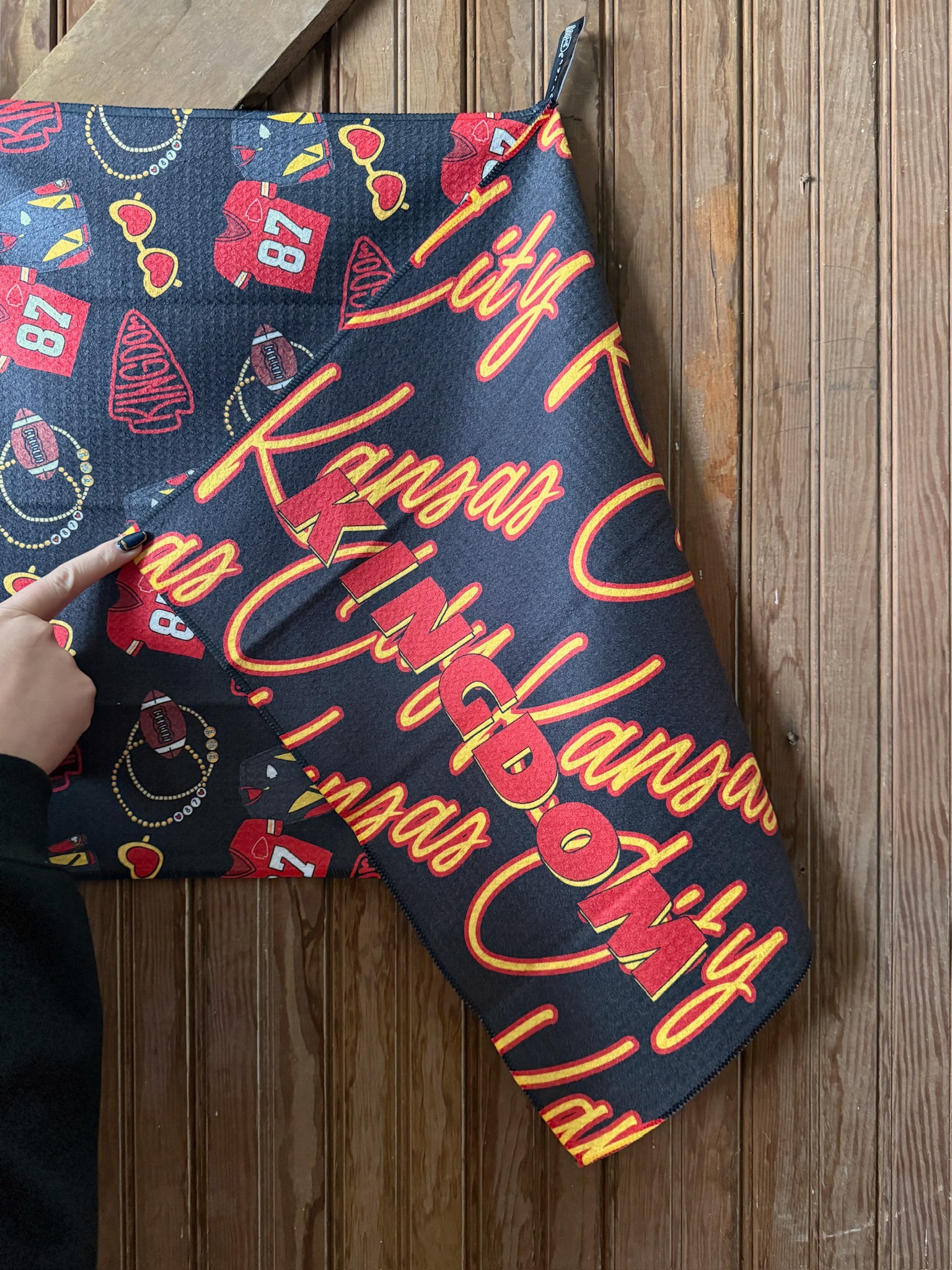 Close-up of a black microfiber tea towel featuring Kansas City Kingdom text in red and gold script with footballs, jerseys, and heart sunglasses, held by hand against a wooden wall.