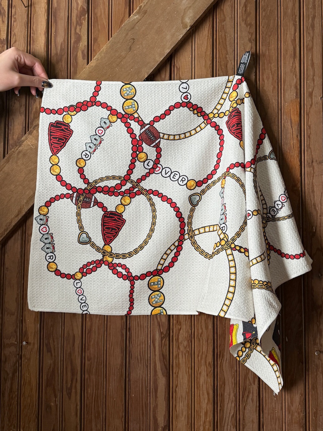 Flat lay of a white microfiber tea towel with red and gold friendship bracelet, football, and heart designs inspired by Taylor Swift and Chiefs Kingdom.