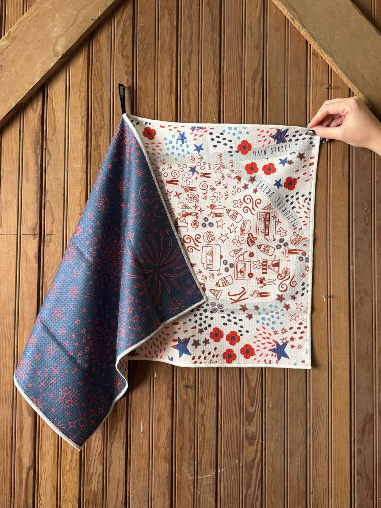 Double-sided red, white, and blue tea towel featuring Kansas City WWI Museum illustrations and patriotic doodles, shown hanging against a wooden wall.