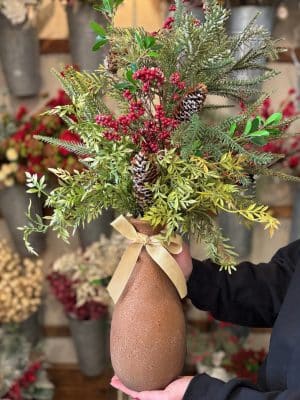 Hand holding a tall clay vase filled with holiday greenery and berries.