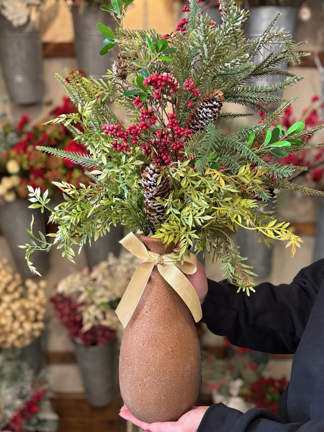 be-made-hays-ks-tall-clay-vase-with-greenery Hand holding a tall clay vase filled with holiday greenery and berries.
