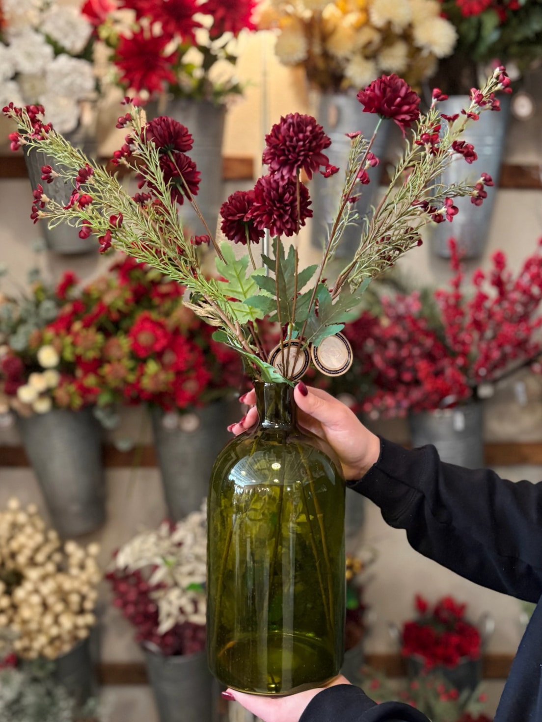 be-made-hays-ks-green-glass-vase-with-florals Hand holding a green glass vase filled with burgundy florals and greenery.