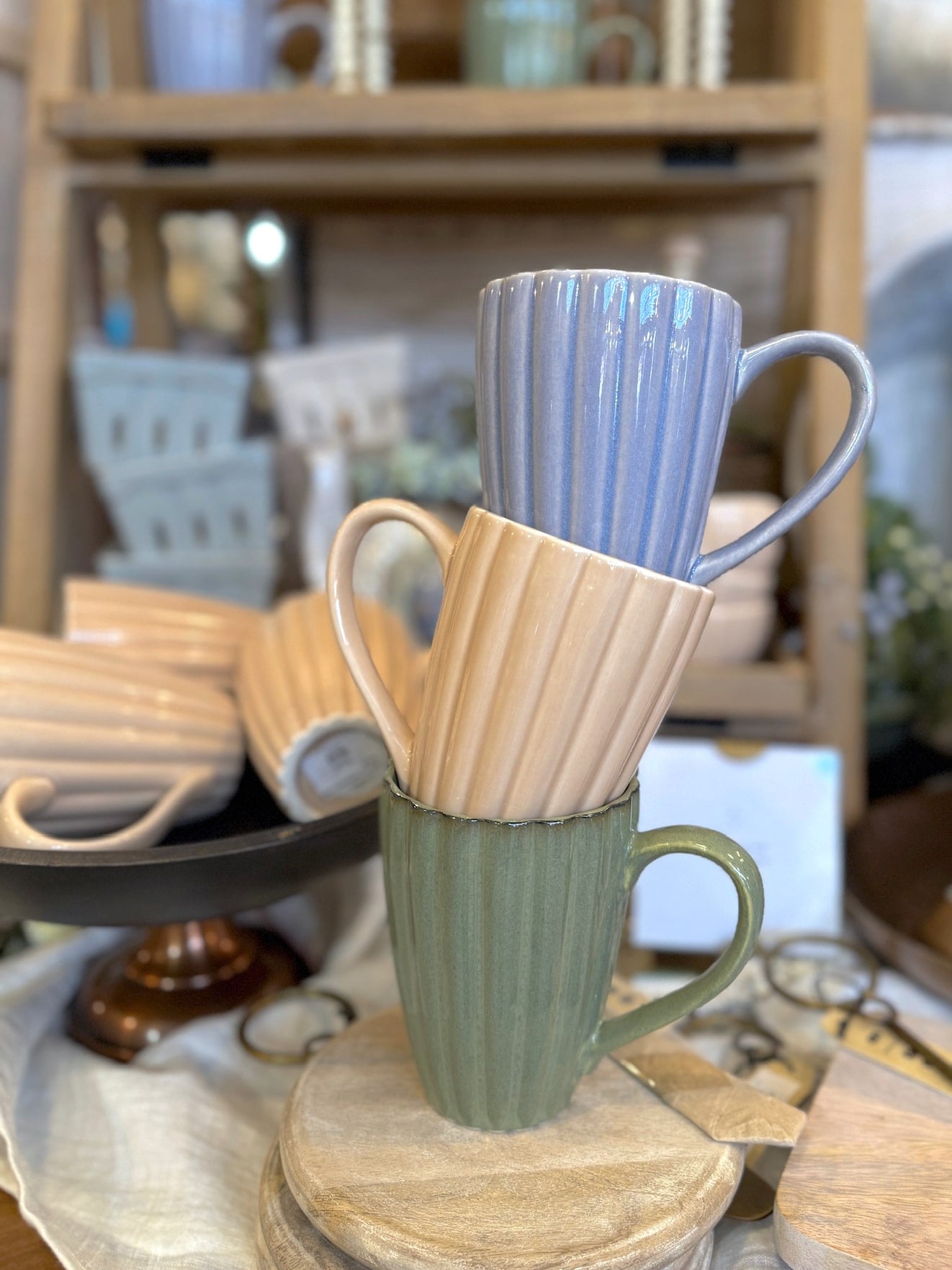 be-made-hays-ks-stoneware-ribbed-mug Stack of three ribbed stoneware mugs in sage green, cream, and light blue with a reactive glaze finish, displayed on wooden risers in a cozy kitchen setting.