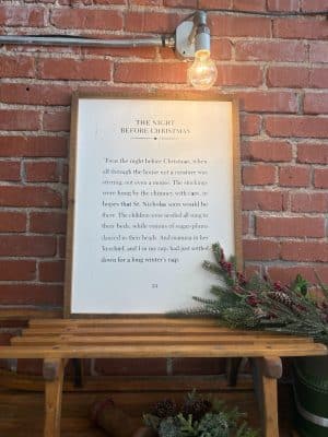 The Night Before Christmas Sign displayed on a brick wall with greenery.