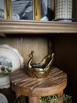 Gold peacock-shaped planter resting on a round rustic wood pedestal, styled on a wooden shelf with books and decorative accents in the background.