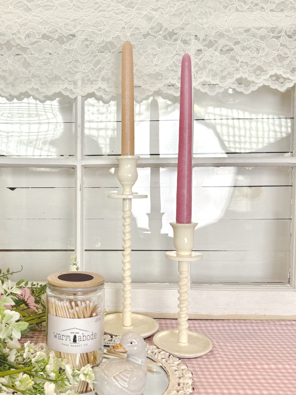 Off-white corkscrew taper candle holders in large and small sizes styled with colored taper candles on a pink gingham table runner