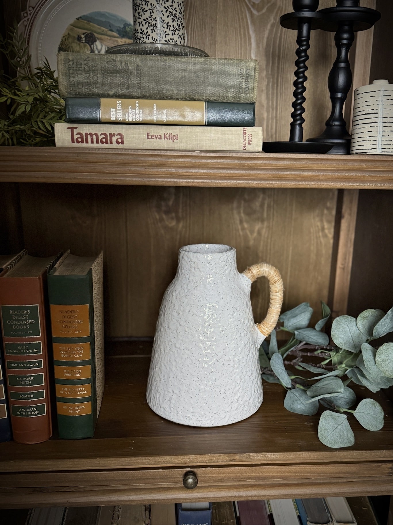 Styled shelf with a small off-white ceramic vase featuring a textured finish and a rattan-wrapped handle, surrounded by vintage books and greenery.