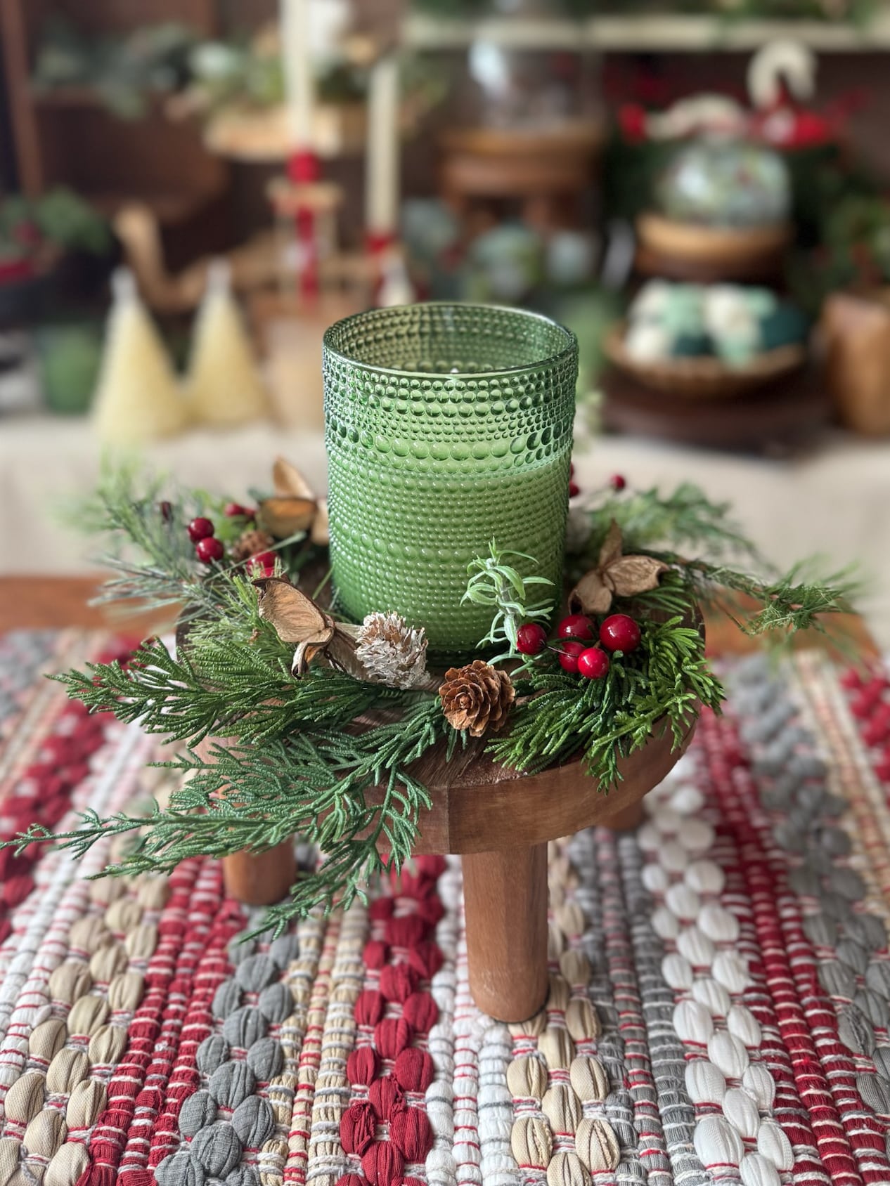 Green dotted flameless candle surrounded by a mini holiday wreath with pine greenery, pinecones, and red berries, displayed on a round wood pedestal over a red and gray Chindi table runner.