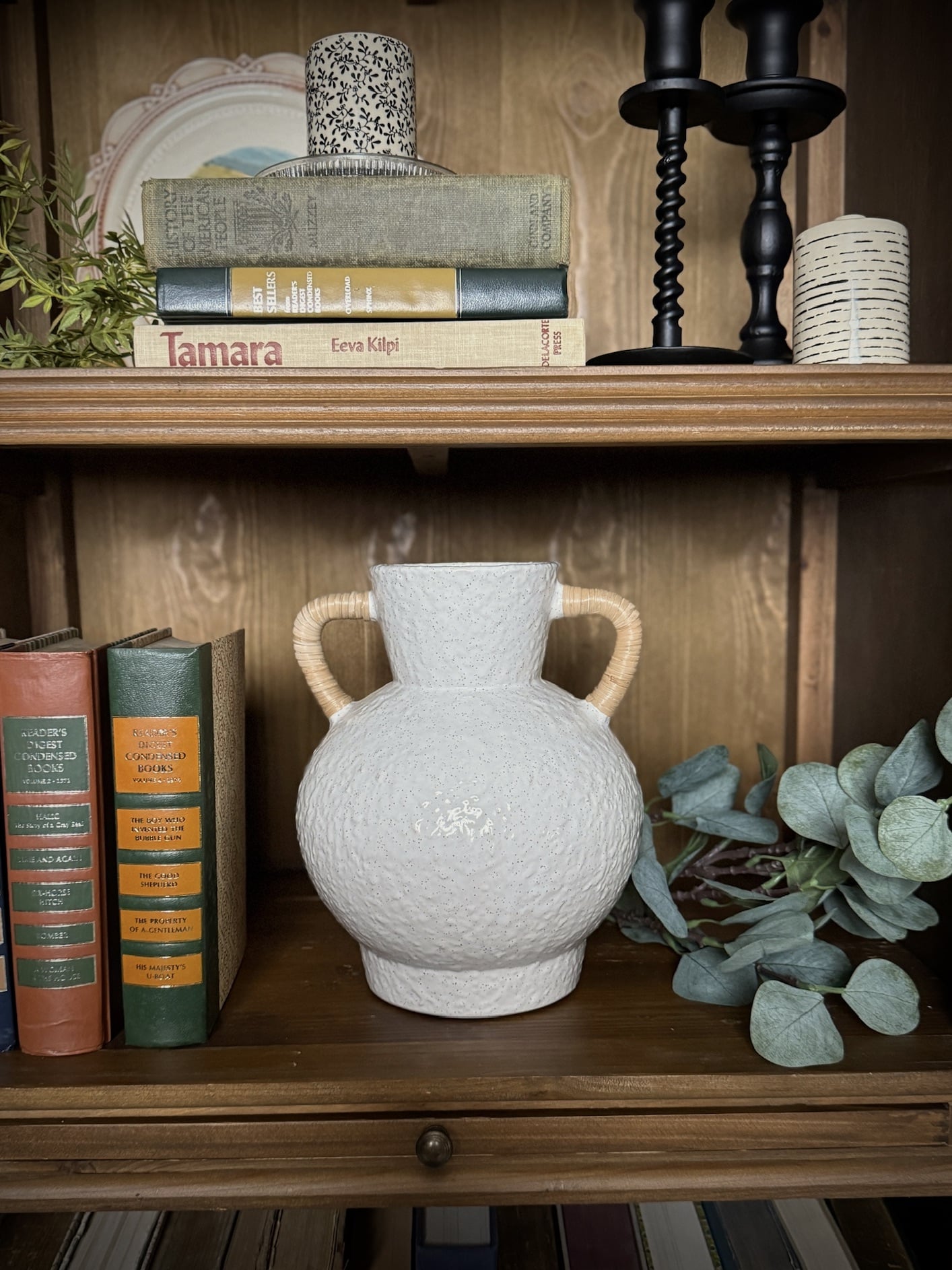 Medium ceramic vase with speckled glaze and two rattan-wrapped handles displayed on a wooden shelf with vintage books and greenery.