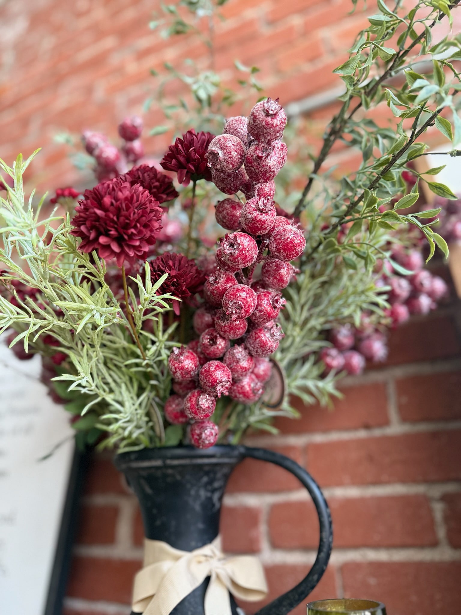 Black metal pitcher filled with Glittered Red Berry Stems and Wispy Green Asparagus Sprays styled against a brick wall for festive holiday décor.