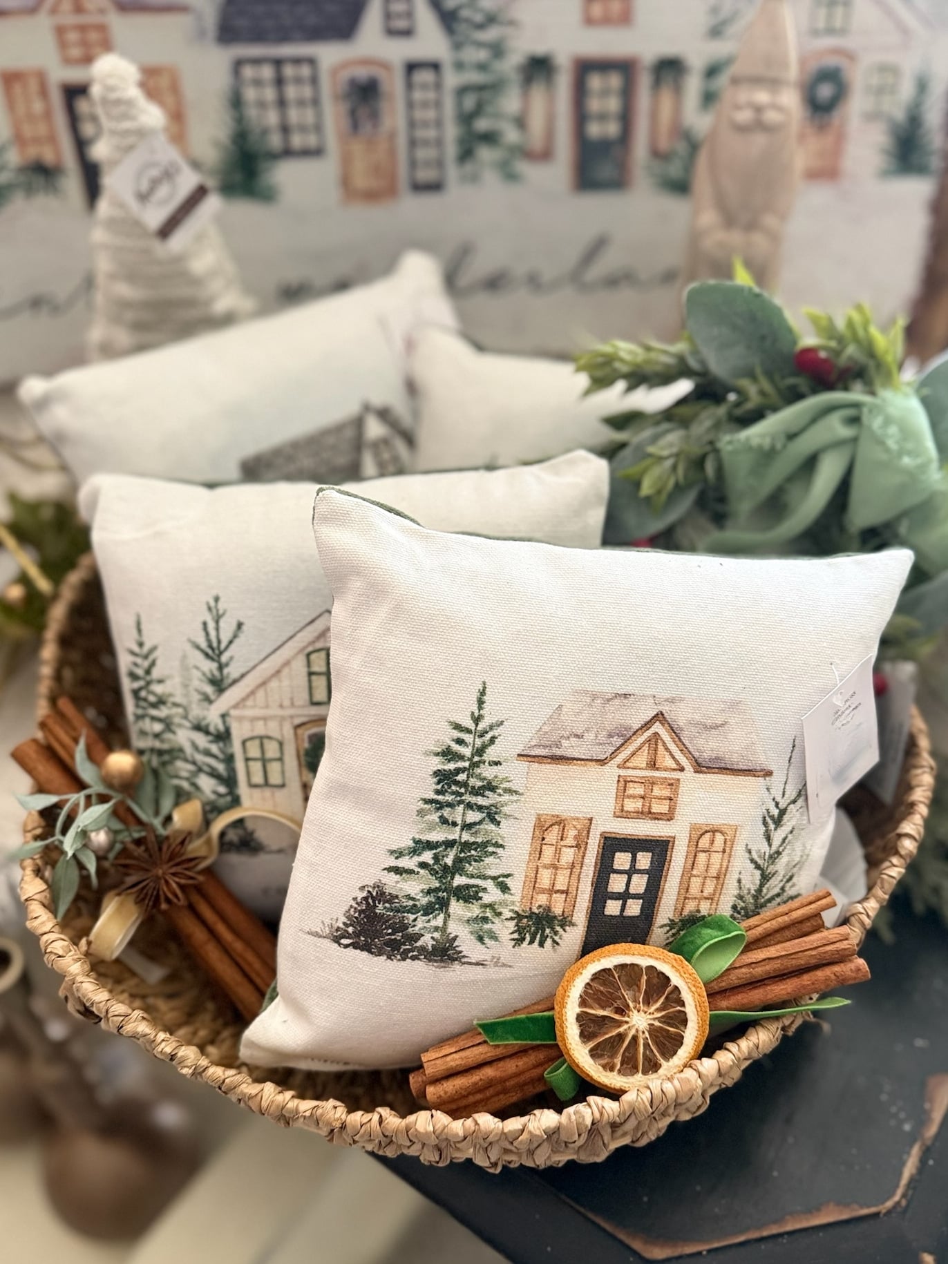 be-made-hays-ks-cozy-winter-cottage-mini-pillows-basket-display Basket filled with Cozy Winter Cottage Mini Pillows featuring illustrated cottages, pine trees, and festive winter details in neutral tones.