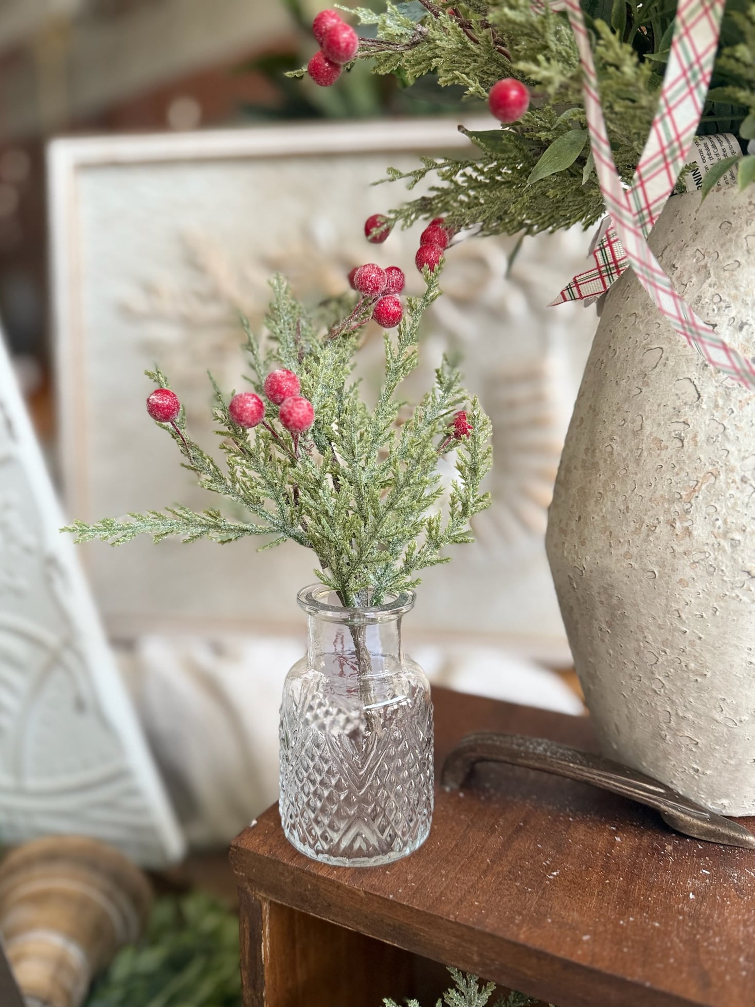 Frosted pine greenery with red glittered berries arranged in a clear pressed glass bud vase on a wooden shelf.