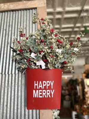 Distressed red metal wall bucket with “Happy Merry” text filled with eucalyptus and red berry floral picks for festive holiday decor.
