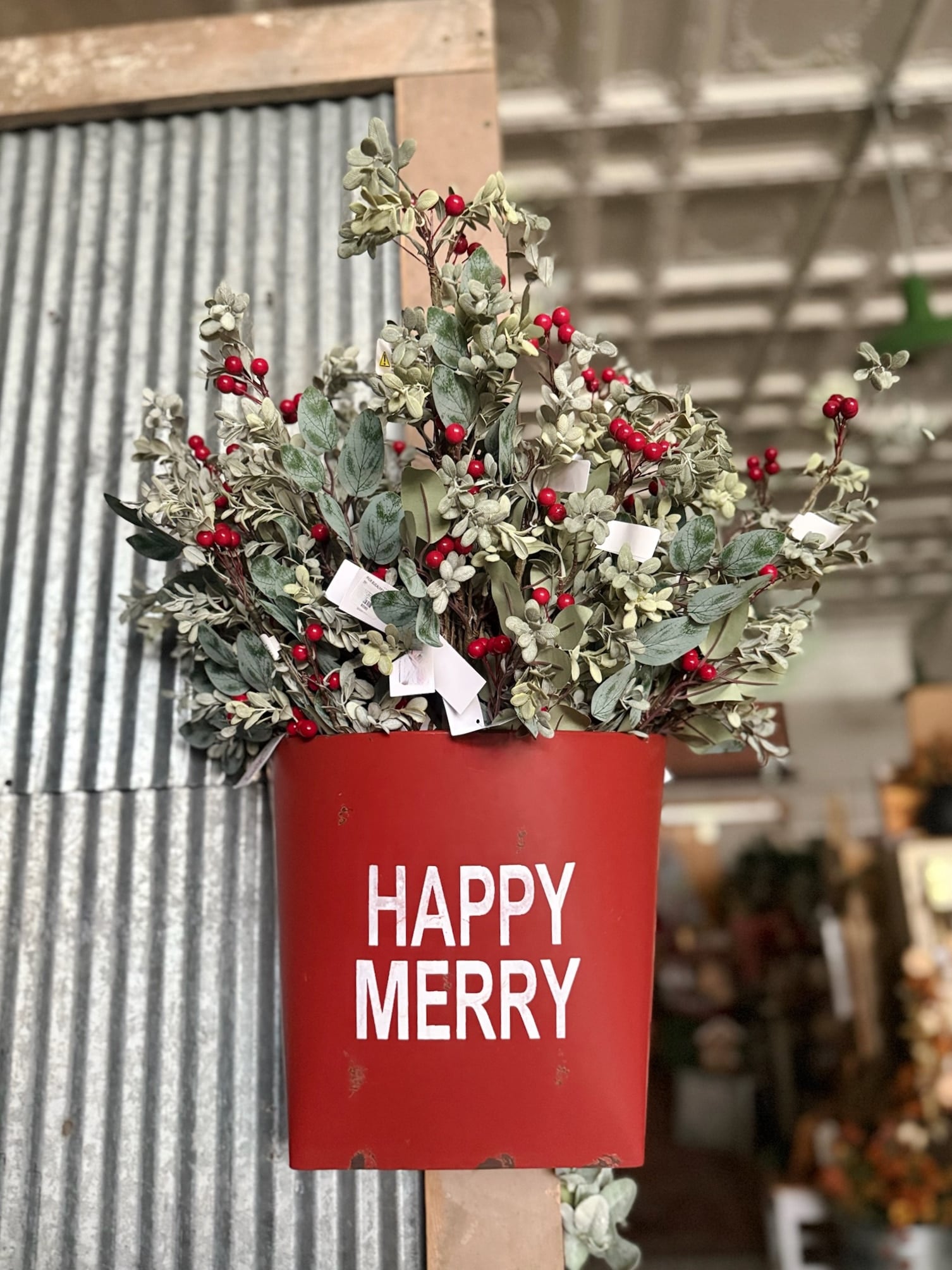 be-made-hays-ks-distressed-red-happy-merry-wall-bucket-with-eucalyptus-and-berries Distressed red metal wall bucket with “Happy Merry” text filled with eucalyptus and red berry floral picks for festive holiday decor.