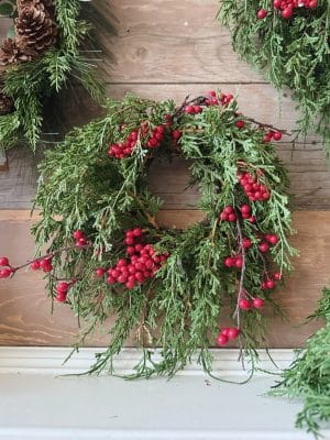 Draping Cedar & Berries Candle Ring with faux cedar greenery and red berries styled against a wooden backdrop.
