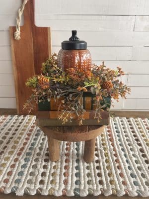 Pumpkin-scented glass candle with black lid surrounded by fall florals on a wood stand over cozy patterned fabric.
