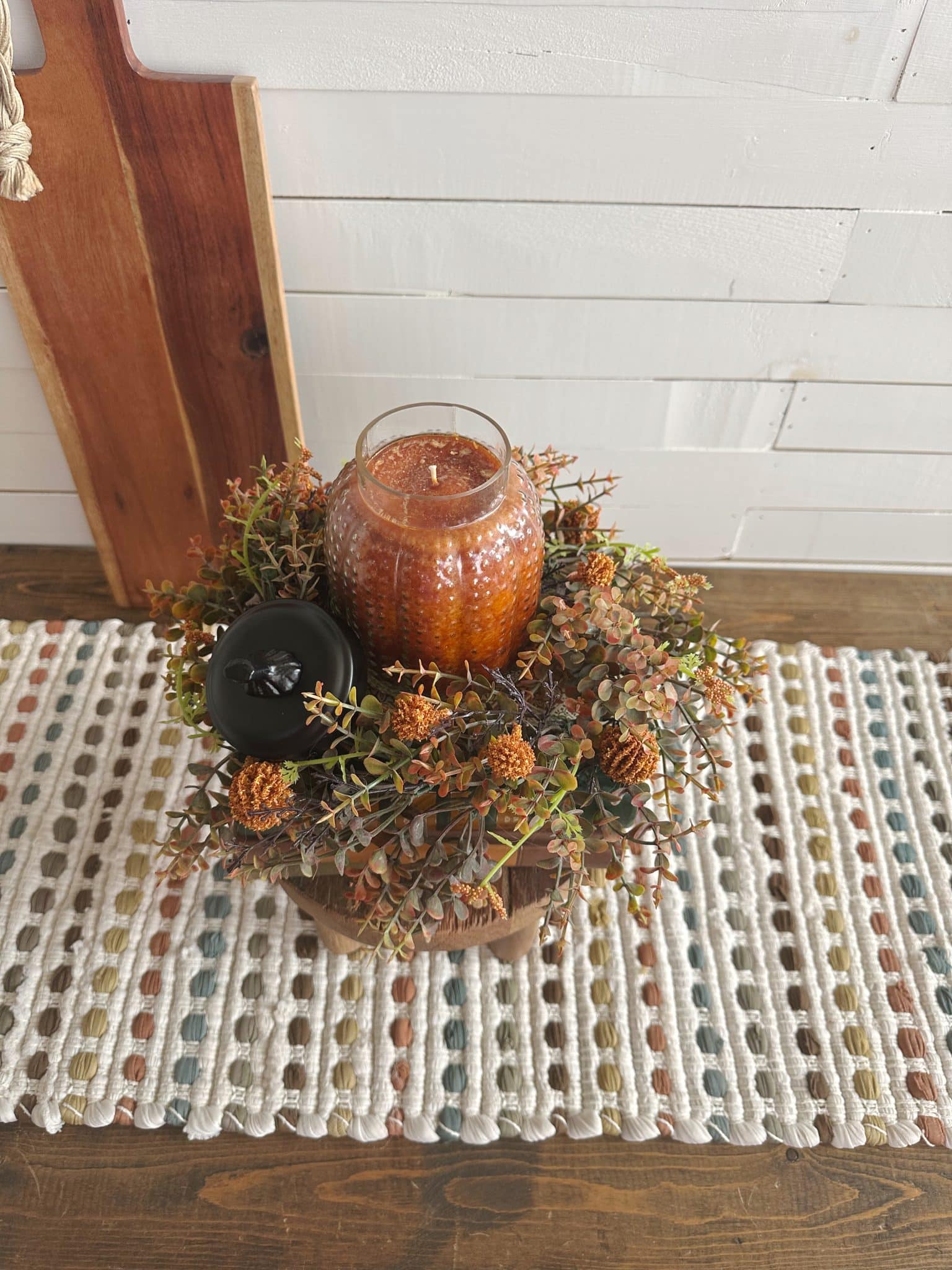 Glass pumpkin candle with fall wreath and black pumpkin accent, styled on a wood pedestal over a textured runner.