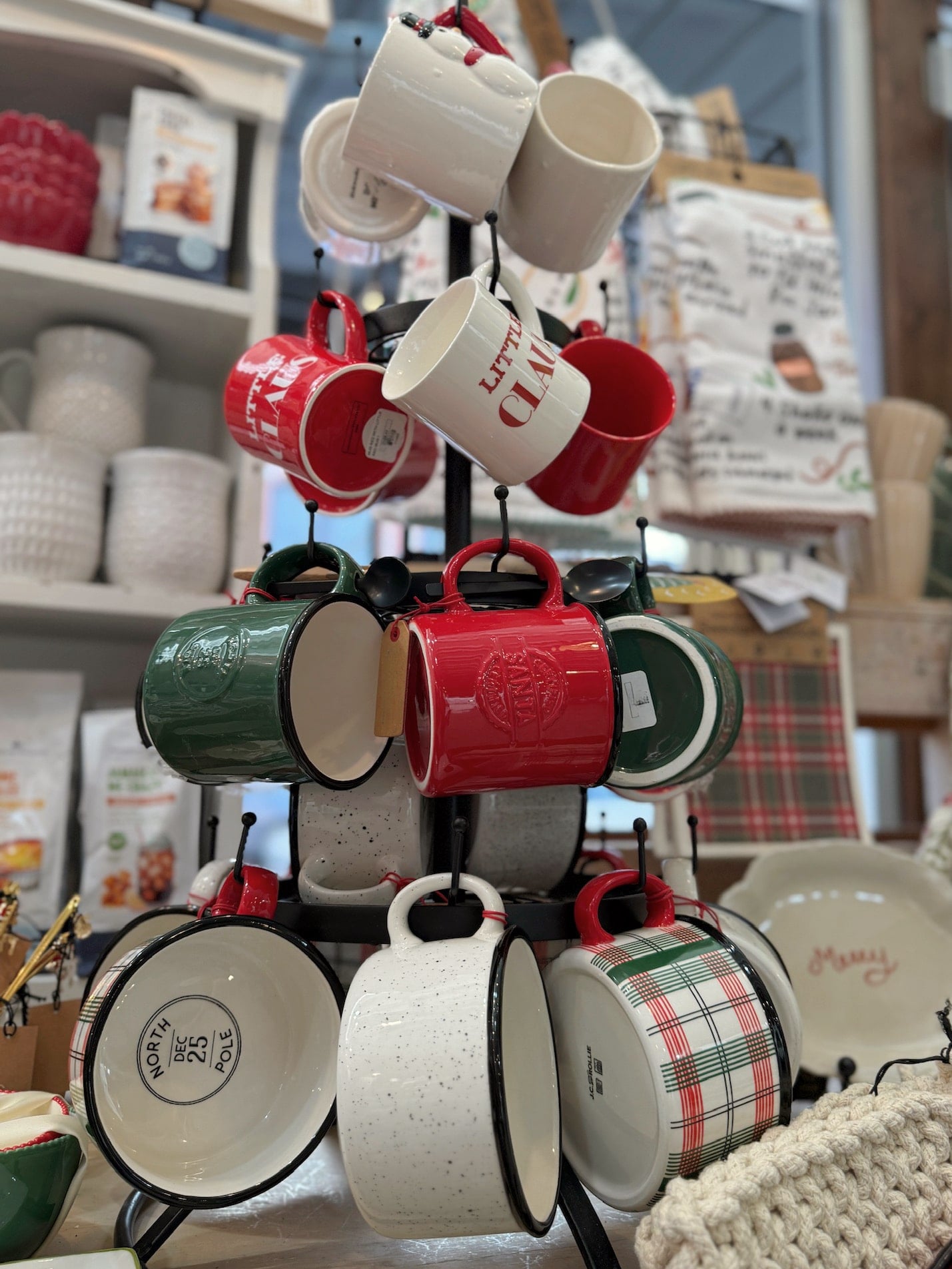 Christmas mug display on a black metal mug tree featuring the White & Black Speckled Hibernate Mug, Christmas Plaid North Pole Mug, Santa Certified Green Mug with Spoon, and Santa Certified Red Mug with Spoon.