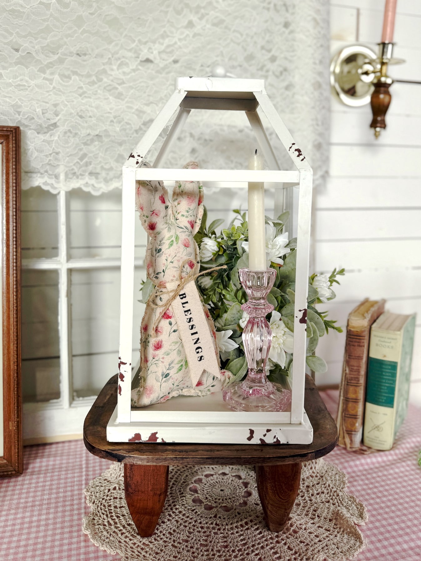 Pink glass taper candle holder styled inside a distressed white metal lantern with floral bunny decor and greenery on a wood stool