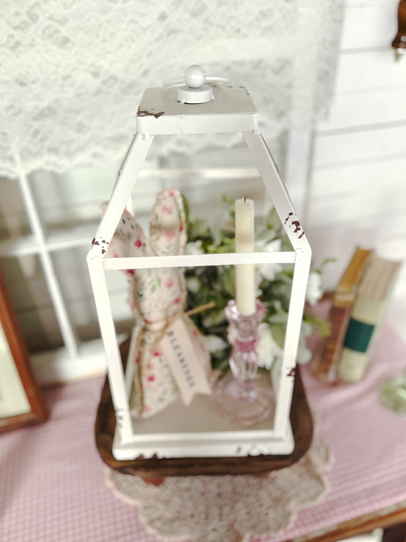 Distressed white metal open frame lantern styled on a wooden riser with pink glass taper candle holder, fabric bunny, and greenery.