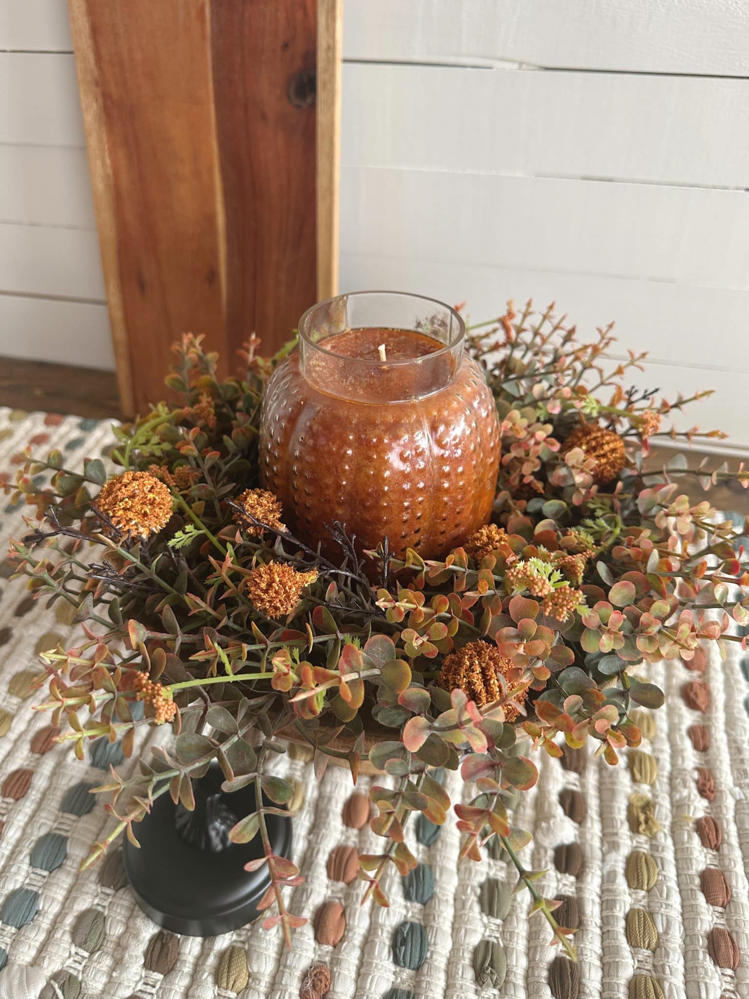 Large glass pumpkin candle surrounded by orange faux blooms and greenery in a pedestal bowl