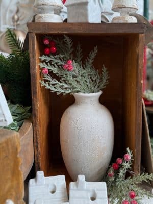 Rustic white vase styled in a wooden crate with glittered pine greenery and red berries for a festive holiday display.