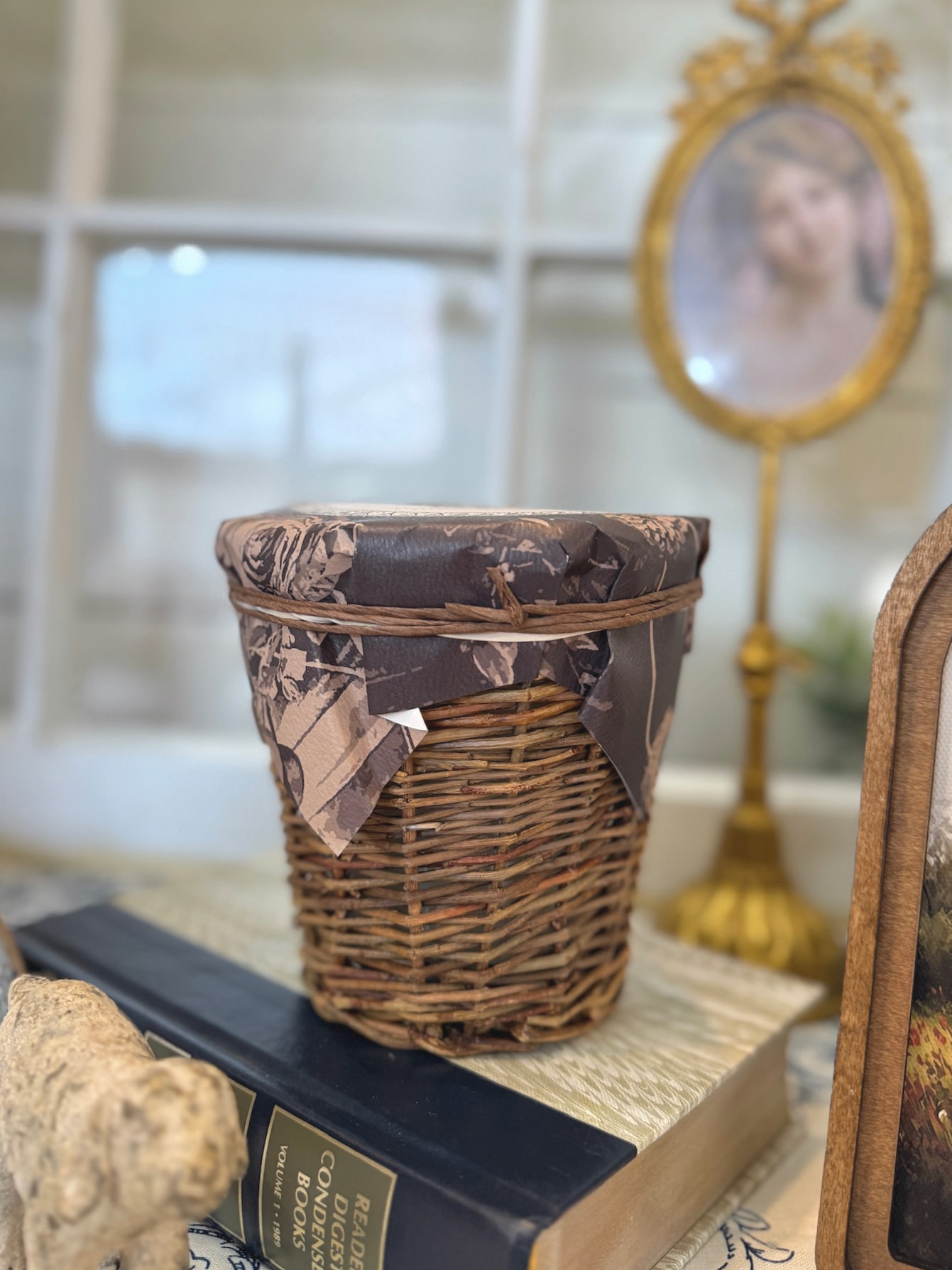 Wicker candle in woven natural basket vessel with paper-wrapped lid styled on stacked books in vintage-inspired setting