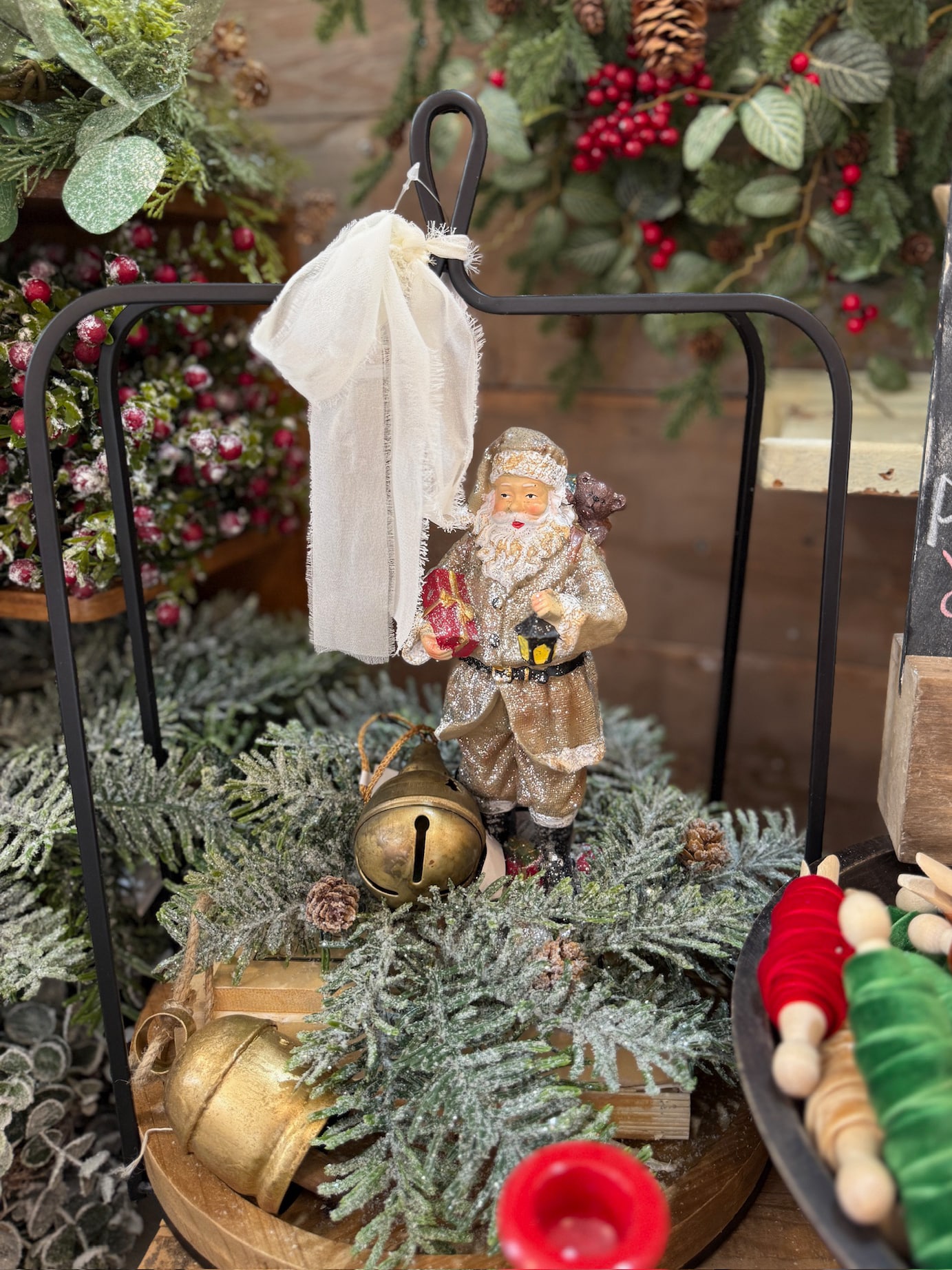 Vintage Santa figurine with lantern styled inside a black open frame lantern on frosted hemlock greenery with gold bells and ribbon.