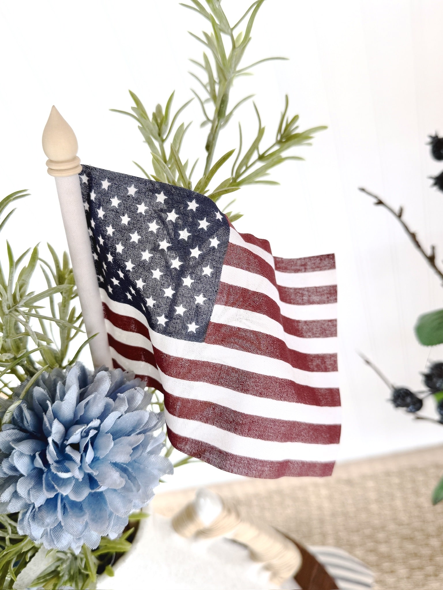 Small American flag on a wooden stick tucked into greenery with a light blue faux flower.