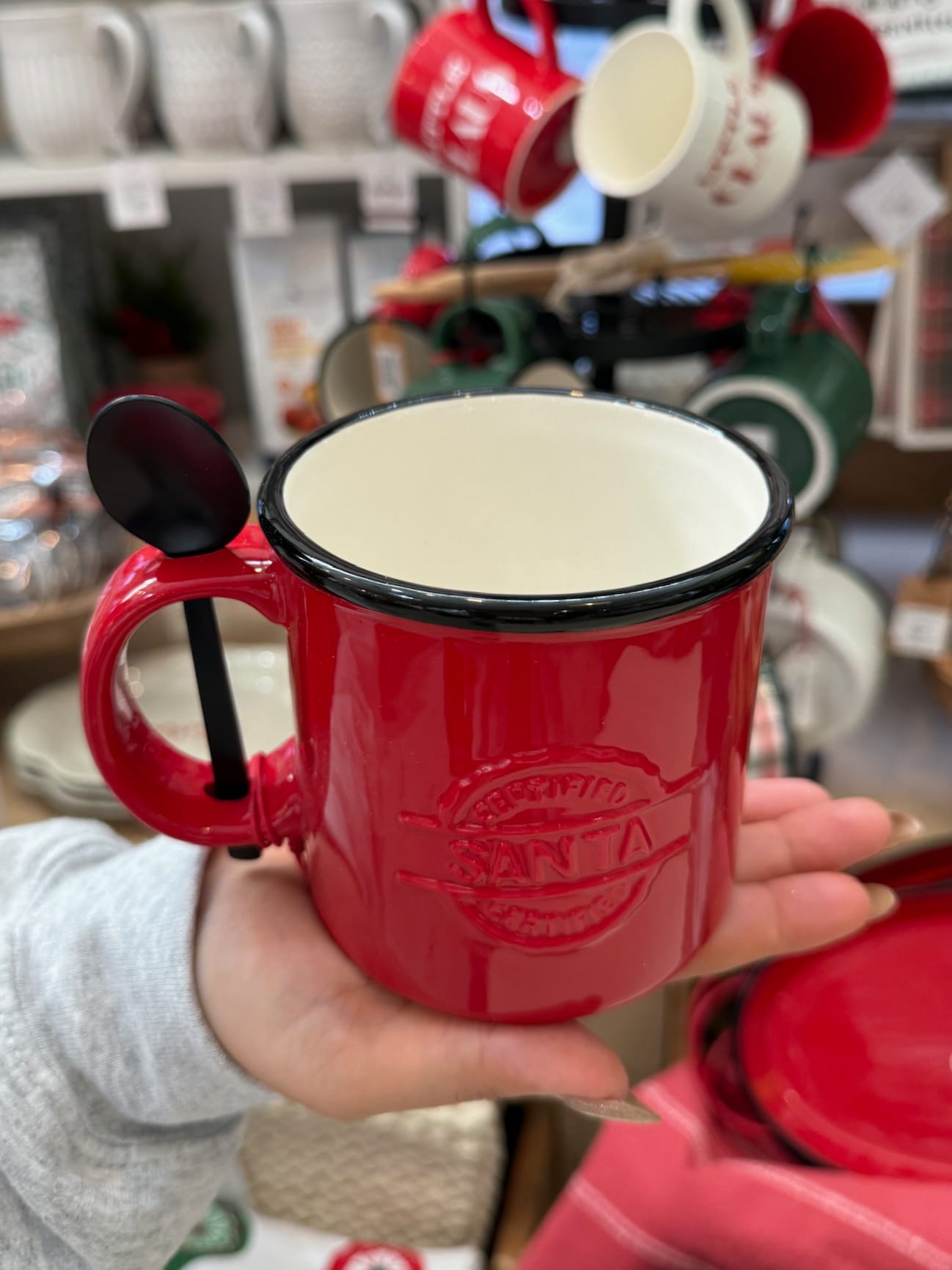 Red “Santa Certified” ceramic mug with black rim and spoon held in the handle, shown in a holiday shop display.