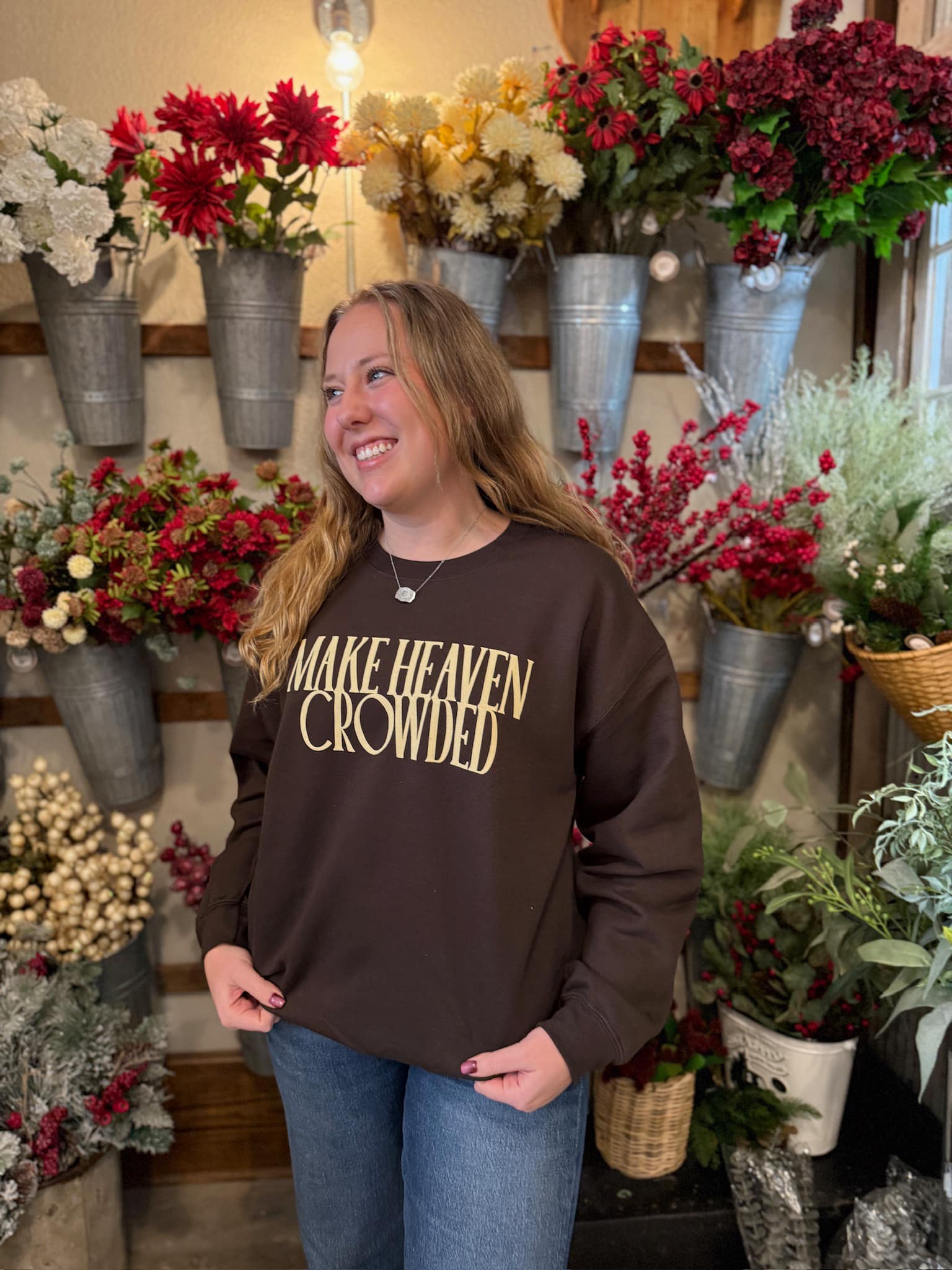 Woman wearing the Make Heaven Crowded Crewneck, smiling in front of floral décor.