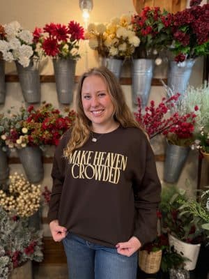 Woman smiling and wearing the Make Heaven Crowded Crewneck in a floral shop setting, showing the relaxed unisex fit and chocolate brown color.