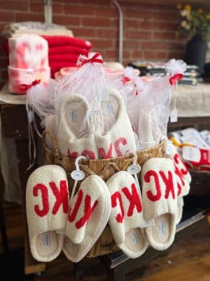White sherpa-style KC slippers with bold red lettering displayed in a basket at Be Made Mercantile.