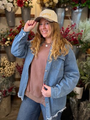 Model in a washed blue denim jacket, brown terry crewneck, and natural trucker hat with gold Tigers embroidery and leopard print bill.